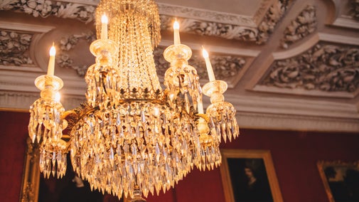 Close-up of an ornate crystal chandelier with candle-style lights hanging from a richly decorated ceiling, illuminated against a backdrop of red walls and framed portraits